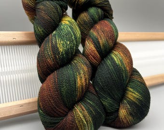 Può includere: Due matasse di filato tinto a mano nei toni del verde, marrone e giallo. Il filato è attorcigliato a forma di matassa, con un attrezzo per maglieria in legno sullo sfondo. Il filato ha un motivo a colori variegato.
