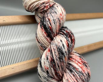 May include: A skein of speckled yarn in cream, black, and red, resting on a wooden weaving loom. The yarn is tightly wound, showcasing a mix of colors and textures. The loom has a white woven surface.