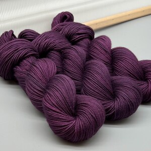 May include: Several skeins of deep purple yarn, tightly wound and displayed on a light grey surface. The yarn has a rich, solid colour and a subtle sheen. A wooden knitting tool is visible in the background, suggesting a crafting theme.