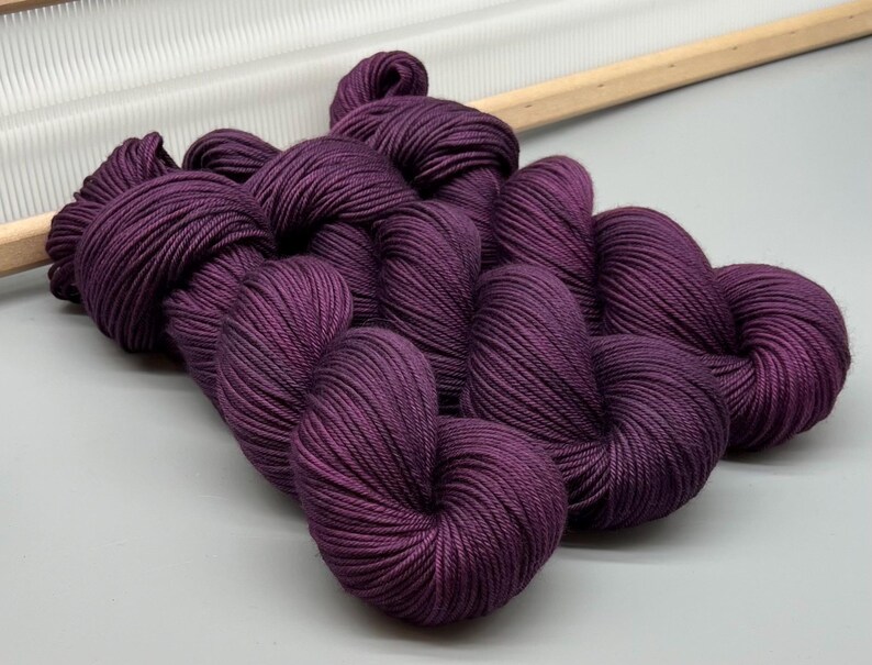 May include: Several skeins of deep purple yarn, tightly wound and displayed on a light grey surface. The yarn has a rich, solid colour and a subtle sheen. A wooden knitting tool is visible in the background, suggesting a crafting theme.