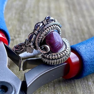 Puede incluir: Un anillo de alambre plateado envuelto con una piedra preciosa púrpura y piedras púrpuras más pequeñas. El anillo está sobre un par de alicates.
