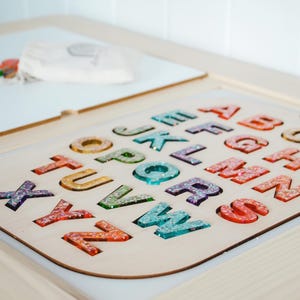 K&ouml;nnte beinhalten: Holz-Alphabet-Puzzle mit bunten, glitzergef&uuml;llten Buchstaben. Die Buchstaben sind in einer hellen Holzbasis angeordnet. Im Hintergrund sind eine wei&szlig;e Oberfl&auml;che und andere farbige Formen zu sehen.