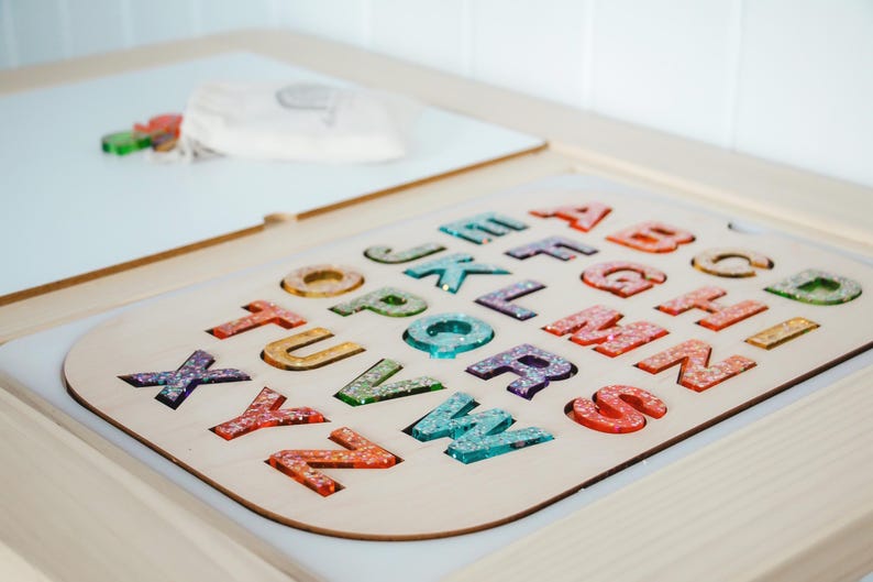 K&ouml;nnte beinhalten: Holz-Alphabet-Puzzle mit bunten, glitzergef&uuml;llten Buchstaben. Die Buchstaben sind in einer hellen Holzbasis angeordnet. Im Hintergrund sind eine wei&szlig;e Oberfl&auml;che und andere farbige Formen zu sehen.