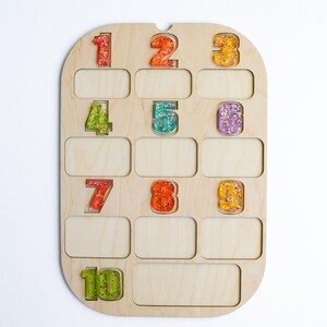 K&ouml;nnte beinhalten: Holzzahlentafel mit zehn farbenfrohen, glitzergef&uuml;llten Zahlen. Die Zahlen sind in verschiedenen Farben, darunter Rot, Orange, Gelb, Gr&uuml;n und Lila. Das Brett hat zehn rechteckige Schlitze unter den Zahlen.