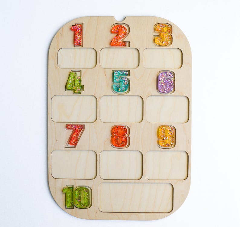 K&ouml;nnte beinhalten: Holzzahlentafel mit zehn farbenfrohen, glitzergef&uuml;llten Zahlen. Die Zahlen sind in verschiedenen Farben, darunter Rot, Orange, Gelb, Gr&uuml;n und Lila. Das Brett hat zehn rechteckige Schlitze unter den Zahlen.