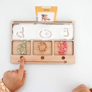 May include: Wooden educational toy with the word "dog" written in sand. The letters "d", "o", and "g" are filled with colourful glitter. A card with a picture of a dog is placed above the letters. A child's finger is pointing at the letter "d".