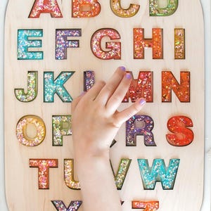 Peut inclure: Puzzle alphabet en bois avec des lettres remplies de paillettes colorées. Les lettres de A à Z sont découpées dans un tableau en bois clair. Une main d'enfant interagit avec la lettre M. Jouet éducatif pour enfants.
