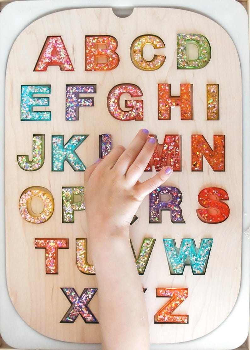 K&ouml;nnte beinhalten: Holz-Alphabet-Puzzle mit bunten, glitzergef&uuml;llten Buchstaben. Die Buchstaben A bis Z sind aus einer hellen Holzplatte ausgeschnitten. Eine Kinderhand interagiert mit dem Buchstaben M. Lernspielzeug f&uuml;r Kinder.