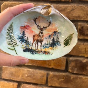 Puede incluir: Un plato de concha decorativo con una escena pintada de un ciervo en un bosque con montañas al fondo. El interior de la concha está pintado en verde pálido con dos hojas de helecho a los lados y un borde pintado en color dorado.