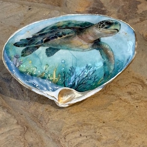 May include: A decorative oyster shell dish featuring a hand-painted scene of a sea turtle swimming underwater. The artwork displays shades of blue and green with coral and seaweed details. The shell has a smooth, glossy finish and a gold-painted rim.