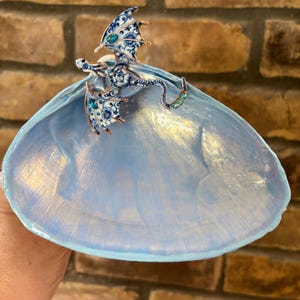 May include: A large, light blue painted seashell dish featuring a decorative metal dragon figurine attached to the top. The dragon has white and blue floral patterns, copper-colored edges, and small teal gemstone accents on its wings and tail.