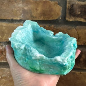 May include: A small, bowl-shaped object with a textured, uneven rim resembling a rocky formation. The surface features a gradient of colors, transitioning from deep green at the base to bright turquoise and white along the top edges.