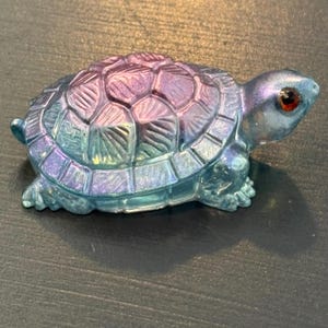 May include: A small turtle figurine featuring a shimmering iridescent finish that shifts between shades of blue, purple, and teal. The shell has a textured, segmented pattern, and the head includes a single, prominent, dark red eye.