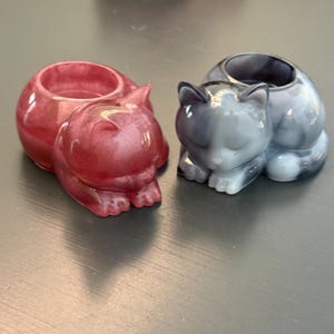 May include: Two cat-shaped holders made of resin. One holder is a shimmering, translucent deep red, while the other is a marbled grey and white. Both feature a rounded, hollow opening on the back, sculpted in the form of a resting feline.