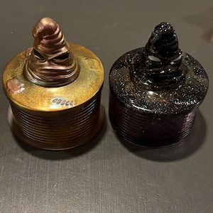 May include: Two cylindrical containers with lids, each featuring a sculpted, pointed hat-like topper with facial features. One container is metallic bronze with a glittery finish, and the other is black with a dense, multi-colored glitter effect.
