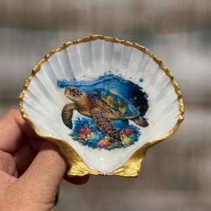 May include: A white scallop shell dish featuring a detailed illustration of a sea turtle swimming above colorful coral reefs. The interior of the shell is white with a glossy finish, while the scalloped edges are painted with a metallic gold trim.