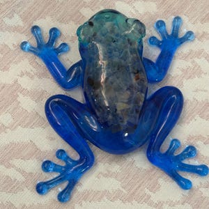 Puede incluir: Una figura de rana de cristal azul translúcido con el dorso hueco relleno de pequeñas piedras de colores. La rana tiene un acabado liso y brillante en las extremidades y la cabeza, con un aspecto texturizado creado por las piedras.