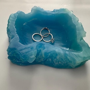 May include: A blue, translucent, wave-shaped resin dish with an irregular, textured rim. Three silver-toned metal rings are resting inside the basin of the dish. The color transitions from a lighter, bright blue to a deeper, darker teal shade.