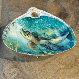 May include: A decorative clam shell dish featuring a detailed illustration of a sea turtle swimming in turquoise water. The shell has a gold-painted rim and a glossy finish, with small bubbles and aquatic plant life depicted in the background.