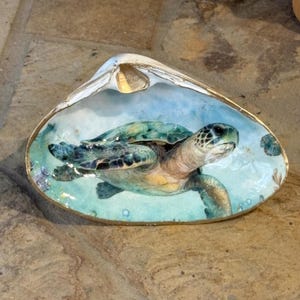 May include: A large, open clam shell featuring a detailed, hand-painted illustration of a sea turtle swimming in light blue water. The interior of the shell is painted in shades of turquoise and white, with a metallic gold trim along the outer edge.