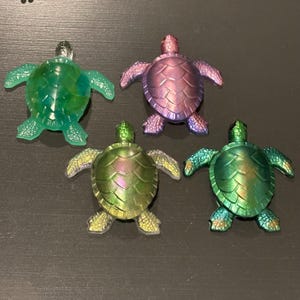 May include: Four turtle figurines arranged in a group. One is translucent green, while the other three feature iridescent finishes in shades of purple, metallic green, and a shifting green-gold. Each turtle has a detailed shell pattern.