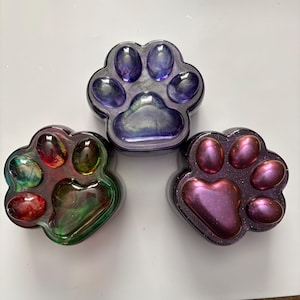 May include: Three paw-shaped resin containers with smooth, glossy surfaces. One is purple with a pearlescent finish, one is green and red with a marbled effect, and one is a deep metallic magenta with subtle glitter throughout the material.