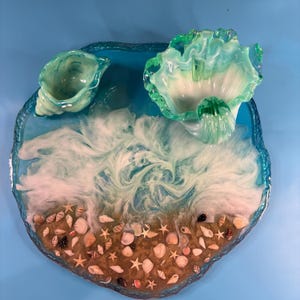 Puede incluir: Una bandeja circular de resina con un diseño de olas oceánicas en azul y blanco con conchas y estrellas de mar incrustadas. Dos cuencos decorativos de cristal en tonos verde y blanco están colocados sobre la superficie, imitando conchas.