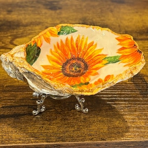 May include: A decorative oyster shell dish featuring a vibrant sunflower print on the interior with bright yellow and orange petals and green leaves. The shell is supported by a small, ornate silver-toned metal stand with three curved legs.
