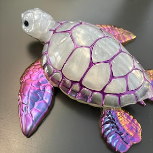 May include: A sea turtle figurine featuring a pearlescent white shell with deep purple outlines. The head is a matching pearlescent white with a dark eye. The four flippers display a vibrant, iridescent gradient shifting between purple, pink, and gold.