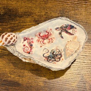 May include: A decorative oyster shell dish featuring a glossy white interior with floral-patterned illustrations of a seahorse, a crab, an octopus, and a sea turtle. The shell edge is trimmed in silver, and two small seashells are attached to the surface.