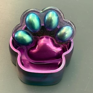 May include: A paw-shaped trinket box with a lid featuring three iridescent teal-to-purple oval pads and a large central purple pad. The base of the container is a deep metallic purple with subtle blue and teal swirls, creating a smooth finish.