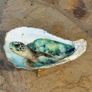 May include: A decorative oyster shell featuring a hand-painted sea turtle in shades of teal, green, and blue. The shell has a smooth white interior, a detailed turtle illustration, and a metallic gold-painted edge, resting on a small gold stand.