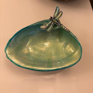 Puede incluir: Un plato decorativo con forma de concha que presenta un acabado brillante e iridiscente en tonos verde azulado, turquesa y dorado. Un adorno de libélula de metal plateado con alas translúcidas está fijado en el borde superior del cuenco.