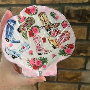 Puede incluir: Una concha de vieira decorativa con un diseño de decoupage de varias botas vaqueras en azul, verde, amarillo y rosa, intercaladas con racimos de rosas rojas y rosas sobre un fondo blanco con un borde festoneado pintado en rosa suave.