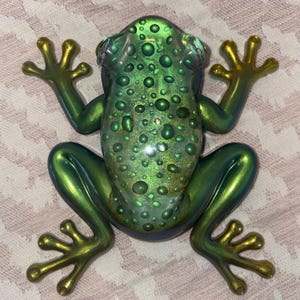 May include: A decorative frog figurine featuring a metallic, iridescent green finish with a textured, bumpy surface. The frog is depicted in a top-down view with four splayed legs, each ending in distinct, rounded toes, showcasing a glossy sheen.
