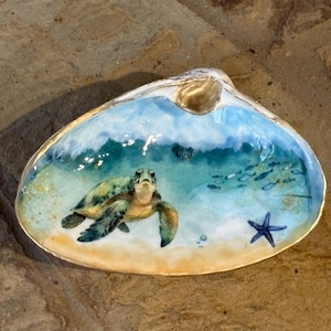 May include: A decorative clam shell featuring a hand-painted underwater scene. The artwork depicts a green sea turtle swimming in clear blue water above a sandy ocean floor, accompanied by a small blue starfish and a school of tiny fish.
