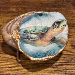 May include: A decorative seashell dish featuring a hand-painted sea turtle swimming in blue water. The interior of the shell is painted with shades of blue and green, while the outer rim is accented with a metallic gold finish and a gold metal stand.