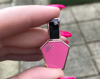 May include: A pink enamel pin shaped like a nail polish bottle with a gold outline and a black cap.