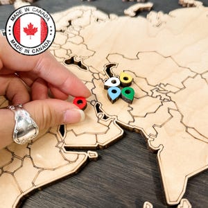 May include: A wooden world map with laser-cut country outlines. A hand places a red marker on the map, near a cluster of colourful markers. The map has a "Made in Canada" stamp. The markers are in various colours: white, yellow, blue, and green.