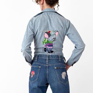 May include: Light blue denim jacket with an embroidered cartoon mouse on the back, paired with flared blue jeans. The jeans feature patches on the back pockets and are worn with red heeled boots. A vintage, retro style.