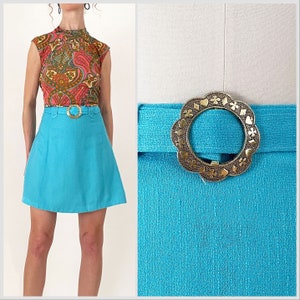 May include: A turquoise blue mini skirt with a gold-toned buckle belt featuring a flower-shaped design. The skirt is made of a textured fabric.