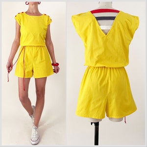 May include: A yellow jumpsuit with a V-neck and short sleeves. The jumpsuit has a drawstring waist and a red drawstring. The jumpsuit is made of a lightweight fabric.