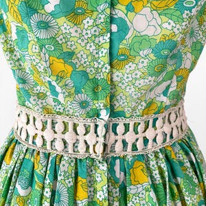 May include: A vintage dress featuring a vibrant floral pattern in shades of green, yellow, and white. The dress has a fitted bodice and a flared skirt, with a cream-colored, openwork belt accentuating the waist. The fabric is a lightweight material.