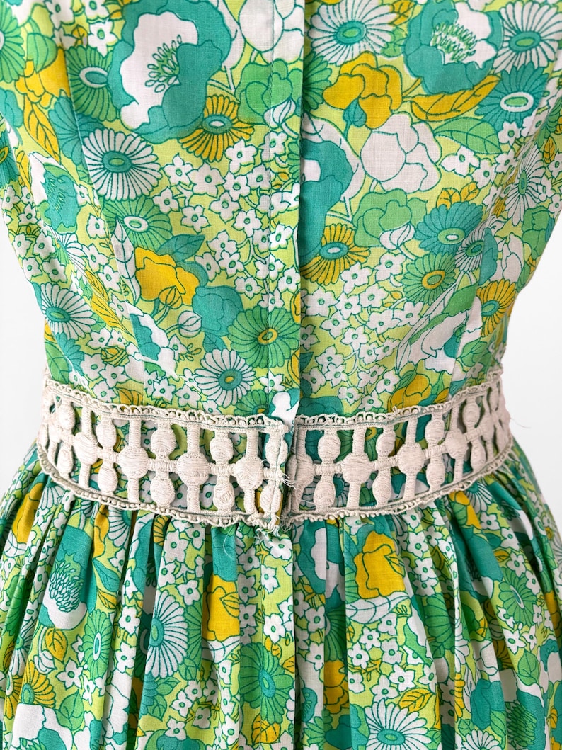 May include: A vintage dress featuring a vibrant floral pattern in shades of green, yellow, and white. The dress has a fitted bodice and a flared skirt, with a cream-colored, openwork belt accentuating the waist. The fabric is a lightweight material.