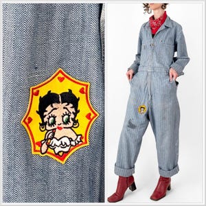 May include: Light blue denim jumpsuit with a Betty Boop patch on the chest and leg. The jumpsuit has a herringbone pattern and is worn with red boots. The Betty Boop patch is yellow with a red border and features the cartoon character.