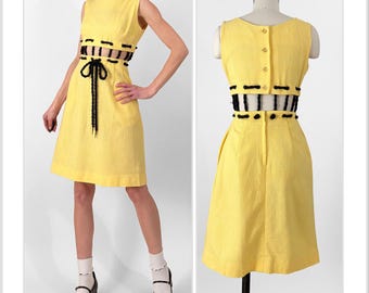 May include: A yellow sleeveless dress with black trim and a black ribbon tie at the waist. The dress has a fitted bodice and a flared skirt.