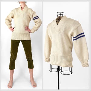 May include: Cream-colored sweater with a V-neck and ribbed cuffs and hem. The sleeves feature two navy blue stripes. The sweater is displayed on a mannequin and worn by a person with green pants.
