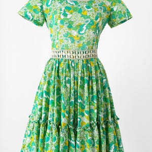 May include: A vintage green floral print dress with a square neckline and short sleeves. The dress features a white crocheted belt and a tiered skirt with ruffles. The fabric has a pattern of white, yellow, and green flowers. The dress is displayed on a mannequin.