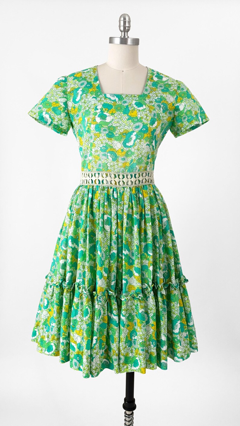 May include: A vintage green floral print dress with a square neckline and short sleeves. The dress features a white crocheted belt and a tiered skirt with ruffles. The fabric has a pattern of white, yellow, and green flowers. The dress is displayed on a mannequin.