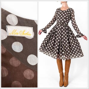 May include: A brown polka dot dress with long sleeves and a flared skirt. The dress features a brown belt and is paired with brown knee-high boots. The label reads "Miss Serbin".